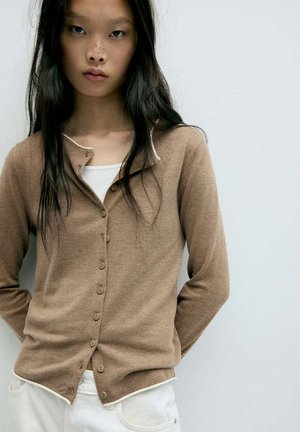Jeune femme aux longs cheveux noirs portant un cardigan marron boutonné par-dessus un haut blanc et un pantalon blanc, debout devant un fond clair uni.