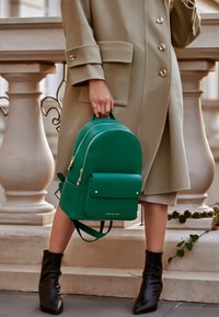 Green leather backpack with gold hardware, rounded shape, front pocket, and adjustable straps, held by a person in a beige coat and black boots.