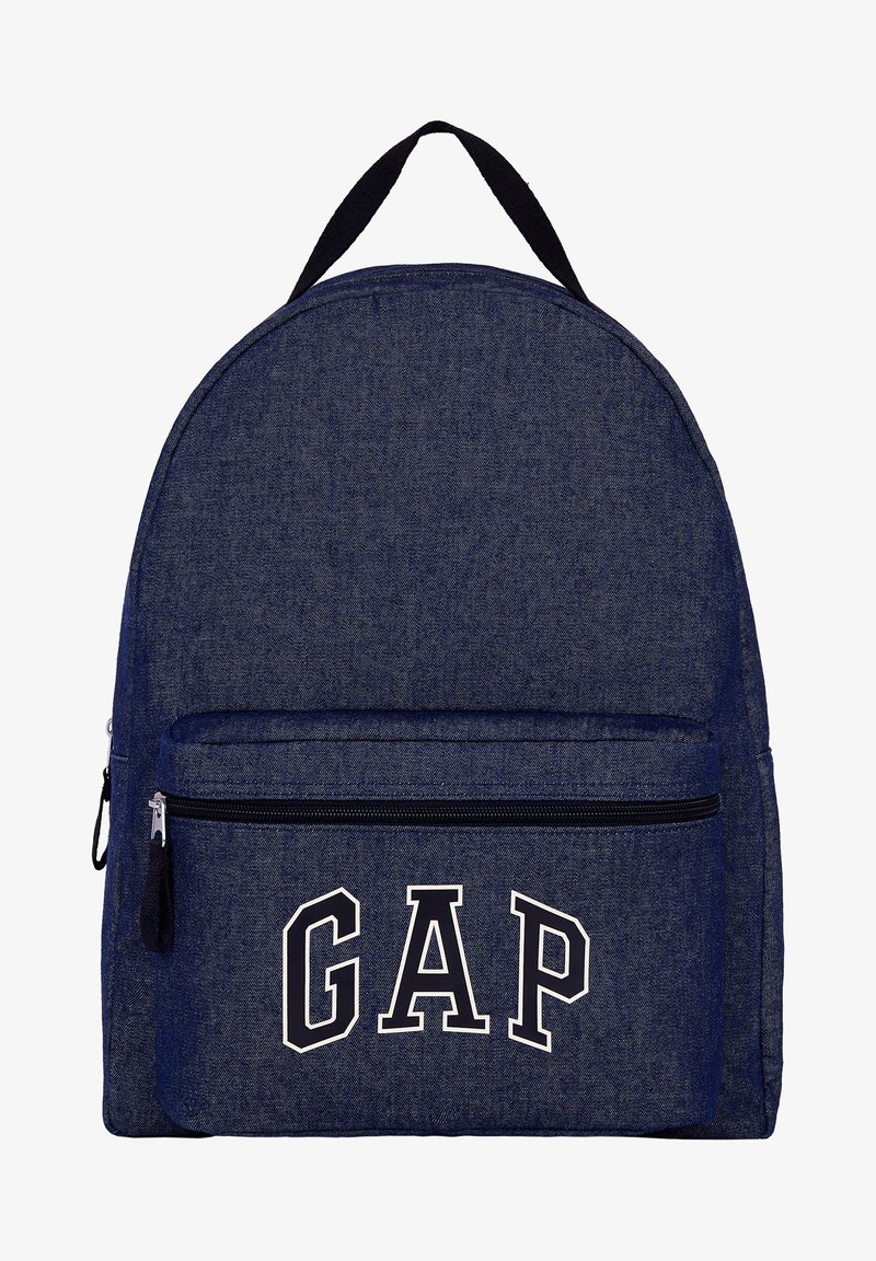 Sac à dos en denim bleu avec fermeture éclair noire et poignée noire, la poche avant affiche de grandes lettres blanches contourées formant GAP.
