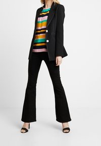 Blazer nero su misura con bottoni bianchi sopra una camicia a righe colorate in rosa, verde e giallo, abbinato a pantaloni neri a zampa d'elefante e tacchi.
