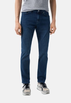 Dark blue denim jeans with a slim fit, featuring standard five-pocket design, button closure, and subtle fading. Paired with neutral-toned sneakers.