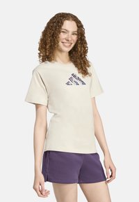 Camiseta de algodón beige de manga corta, con un diseño de logo geométrico en color azul marino en la parte delantera. Combinada con pantalones cortos morados.