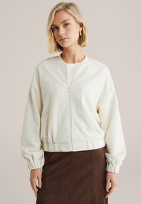 Krémová bomber bunda s textúrou, so zipsom vpredu, rebrovanými manžetami a pásom. Nosená cez biely top, skombinovaná s hnedou sukňou.