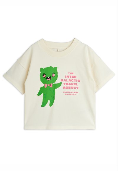 Κοντομάνικο μπλουζάκι εκρού με ένα πράσινο χαμογελαστό εξωγήινο που φοράει ένα ροζ παπιγιόν και κείμενο που γράφει «The Inter Galactic Travel Agency».