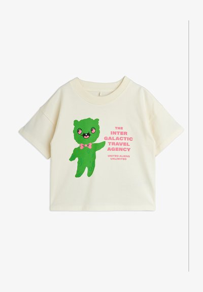 Κοντομάνικο μπλουζάκι εκρού με ένα πράσινο χαμογελαστό εξωγήινο που φοράει ένα ροζ παπιγιόν και κείμενο που γράφει «The Inter Galactic Travel Agency».