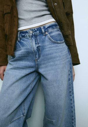 Lysblå denim jeans med en løs pasform, der har en knaplukning, frontlommer og subtil slid. Parret med en grå top og en brun jakke.