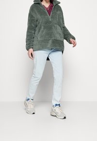 Zielony polarowy półregata z zamkiem błyskawicznym, teksturowana powierzchnia, kieszeń kangurka, w połączeniu z jasnoniebieskimi dżinsami i wielokolorowymi trampkami.