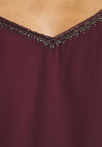 Gros plan sur un tissu bordeaux avec un décolleté en V orné de délicates perles noires et argentées.