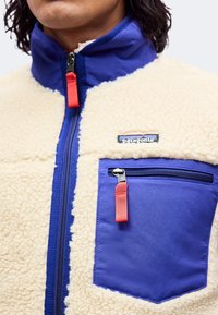 Κοντινή λήψη fleece μπουφάν με κρεμ ύφασμα sherpa, μπλε φερμουάρ και τσέπη στο στήθος, και τα δύο με κόκκινα γάντζους φερμουάρ, και ένα έμβλημα με το λογότυπο Patagonia.