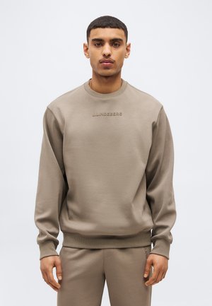 Mladý muž v béžovej mikine s okrúhlym výstrihom a zodpovedajúcich nohaviciach, stojaci pred jednoduchým bielym pozadím.