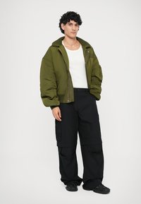 Jeune homme avec des cheveux bouclés portant une veste bomber olive, un débardeur blanc, un pantalon cargo noir et des baskets noires, se tenant devant un fond uni.