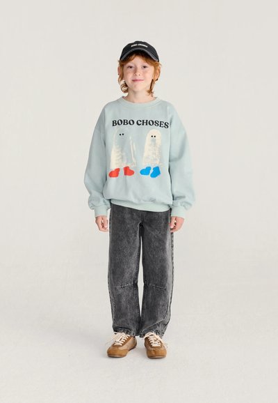 Bambino con capelli rossi indossa un berretto nero, una felpa azzurra con fantasmi dei cartoni animati, jeans grigi e scarpe da ginnastica marroni, in piedi davanti a uno sfondo semplice.