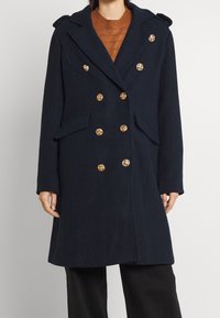Donna che indossa un cappotto blu navy doppiopetto con bottoni dorati sopra un maglione in maglia marrone e pantaloni neri, in piedi su uno sfondo neutro.