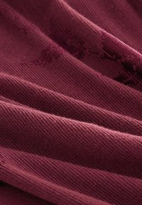 Burgundská tkanina s textúrovaným pleteným vzorom, ktorá obsahuje vrstvené záhyby, ktoré zvýrazňujú jej jemnú, hladkú štruktúru a bohatú farbu.