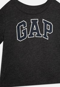 Tamsiai pilkas medvilninio marškinėlių dizainas, puošiamas ryškiai balta ir tamsiai mėlyna "GAP" logo. Apvalus kaklas ir trumpi rankovės, su minkšta, lygiomis tekstūra.