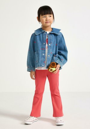 Nuori tyttö sinisessä farkkutakissa, jossa on punaisia pilkkuja, korallinväriset housut, valkoiset tennarit, ja hän pitää kädessään ruskeaa nallekarhuavaimenperää.