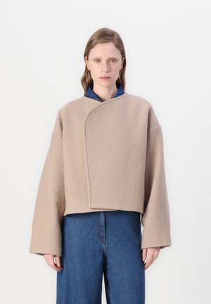 Veste en laine beige, coupée, avec un design croisé, des manches larges et une couture simple le long des bords, portée par-dessus une chemise en denim bleu.