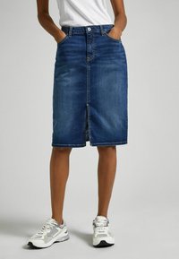 Denim midi skirt in dark blue, featuring front pockets, a central split seam, and a classic button closure. Paired with white and gray sneakers.