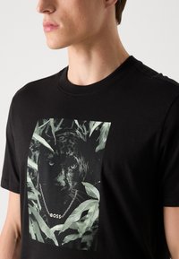 Zwarte katoenen T-shirt met een centraal grafisch ontwerp van het gezicht van een panter, omringd door groene bladeren, met het "BOSS"-logo aan een zilveren ketting.
