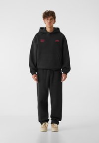 Fekete kapucnis pulcsi és melegítőnadrág szett, elől egy zsebbel, amelyen rózsaszín felirat szerepel: "Vad a szívében." Pamutból készült, laza szabással és állítható kapucnival.