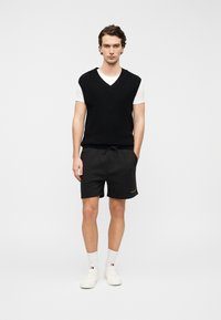 Μαύρο πλεκτό γιλεκό πάνω από λευκό μπλουζάκι, συνδυασμένο με μαύρο σορτς και λευκά αθλητικά παπούτσια. Διαθέτει V-λαιμόκοψη και ζώνη με κορδόνι στη μέση.