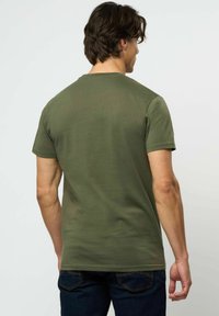 Vīrietis ar tumšiem matiem, ģērbies olīvu zaļā īsroku T-kreklā un tumši zilās džinsās, stāvot ar muguru pret vienveidīgu fonu.