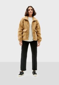 Tan, fluffy fleece jack met een ritssluiting aan de voorkant, twee voorzakken en een opgevouwen kraag. Draagt over een wit topje en zwarte broek. Zwarte sneakers.