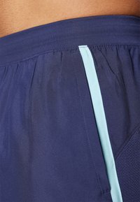 Närbild av midjebandet på blåa träningsshorts med ljusblå vertikalrand och nätpanel på sidan.