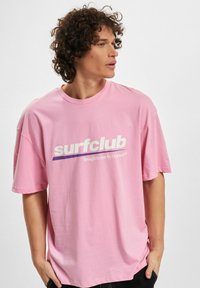 Rosa bomull t-skjorte med kort erm, med "surfclub" logo i hvitt med lilla aksent. Uformell passform med rund hals.