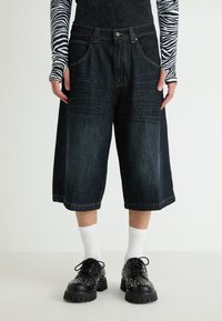 Shorts de mezclilla de color azul oscuro, de pierna ancha y cintura alta, con cinco bolsillos, ligeras decoloraciones y detalles de costura amarilla. Llevados con zapatos negros con tachuelas.