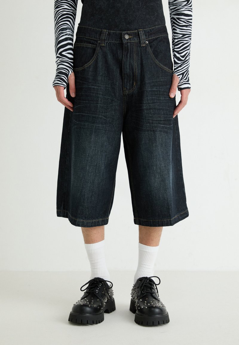 Shorts de mezclilla de color azul oscuro, de pierna ancha y cintura alta, con cinco bolsillos, ligeras decoloraciones y detalles de costura amarilla. Llevados con zapatos negros con tachuelas.