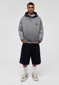 Grijze hoodie met een zwart logo en een gelaagde zwarte capuchon, gecombineerd met donkere shorts en witte sokken. Gedetailleerde sneakers in crème en zwart.