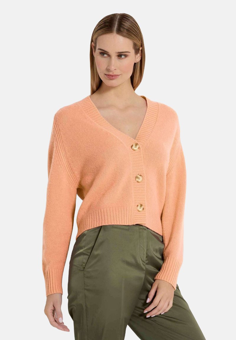 Licht koraalkleurige gebreide cardigan met een V-hals, voorzien van vier gouden knopen aan de voorkant en geribde mouwen, gecombineerd met olijfgroene broek.