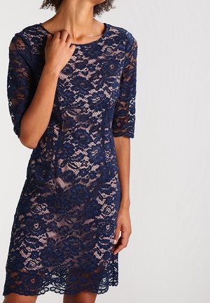 Vrouw met een marineblauwe kanten jurk met bloemenmotief en halve mouwen, staand tegen een effen lichte achtergrond, één hand raakt haar halslijn aan.