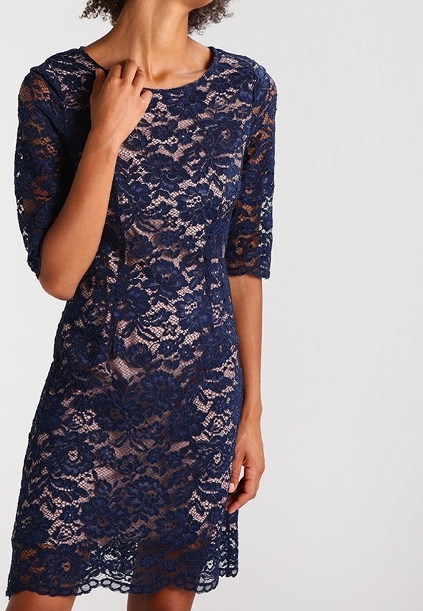 Femme portant une robe en dentelle bleu marine à motifs floraux et manches mi-longues, debout devant un fond uni clair, une main touchant son encolure.