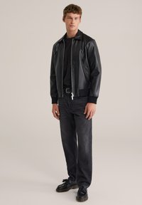 Juoda odinė bombonešio striukė su aptempiamu modeliu, užtrauktuku priekyje ir briaunuotais rankogaliais, derinama su tamsiais džinsais ir juodais batais, stovint neutraliame fone.