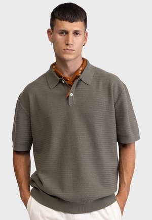 Νεαρός άνδρας που φοράει μπλούζα πόλο με κοντά μανίκια σε ελαιοπράσινο με υφή και καφέ σκουφάκι με σχέδιο, στέκεται με τα χέρια στις τσέπες μπροστά σε απλό φόντο.