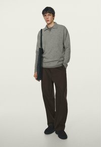 Grå strikket sweater med krave, parret med mørkebrune breddebenbukser. En sort skuldertaske hænger over torsoen.