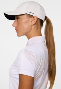 Biała czapka z ciemnym daszkiem, z logo po boku. Koszula w jasnym kolorze z delikatnymi wzorami i standardowym kołnierzykiem.
