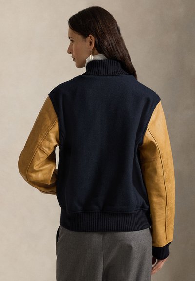 Ναυτική bomber jacket από μίγμα μοκέτας με καφέ δερμάτινα μανίκια, ριμπ μανσέτες και ψηλό γιακά. Μαλακή υφή, τολμηρός χρωματικός συνδυασμός, κομψός σχεδιασμός.