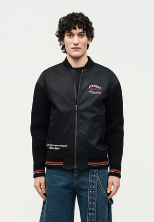 Młody mężczyzna o kręconych włosach, ubrany w czarną kurtkę na zamek z czerwonym i białym napisem oraz niebieskie dżinsy z detalem paska z oczkami.