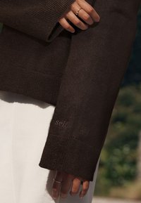 Ruskea neulottu villapaita, jossa on resorireunos ja sana "self" hienovaraisesti kirjailtuna vaaleammalla sävyllä. Lähikuva kädestä, jossa on sormus.