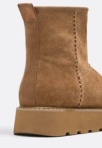 Brązowy zamszowy botek za kostkę z przeszyciami, zaokrąglonym noskiem i grubą, fakturowaną podeszwą na platformie. Detal szwów dodaje atrakcyjności wizualnej.