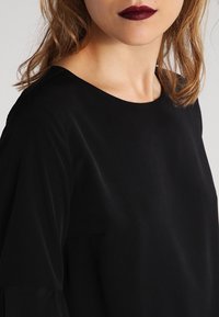 Maglietta nera con scollatura rotonda, tessuto morbido e vestibilità comoda. La texture appare soffice, con elementi di design delicati sulle maniche.