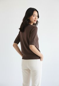 Brązowy krótki sweter z krótkim rękawem o dopasowanym kroju, z prążkowanymi mankietami i obszyciem na dole, zestawiony z białymi spodniami, widoczny od tyłu.