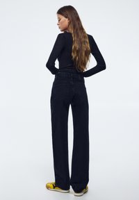 Zwarte wijde jeans met een hoge taille en twee achterzakken, gecombineerd met een aansluitend zwart shirt met lange mouwen en gele sneakers.