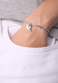 Handgelenk mit silbernem Armband mit herzförmigem Schloss- und Schlüsselanhänger, Hand ruht in der Hosentasche einer weißen Hose, graues Hemd sichtbar.