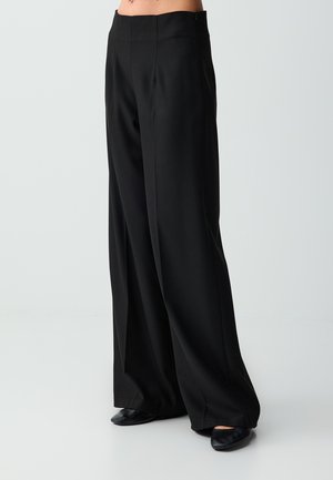 Modell trägt hoch taillierte schwarze weite Hosen kombiniert mit schwarzen spitzen Schuhen vor einem einfarbigen hellen Hintergrund.