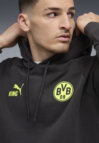 Ung mand justerer en sort hættetrøje med gult Puma-logo, "KING" tekst og BVB 09 fodboldklub emblem, mens han ser til siden.