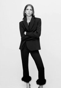 Blazer noir ajusté avec des épaules structurées, associé à un pantalon noir avec des accents en fourrure au niveau des chevilles. Design minimaliste et épuré.